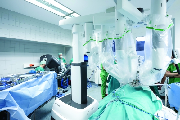 Robotsebészeti műtő setup a daVinci Xi robotkonzollal (bal oldalon, a háttérben) és a transzorálisan dokkolt robotkarokkal (jobb oldalon, az előtérben) MO-MT-archív